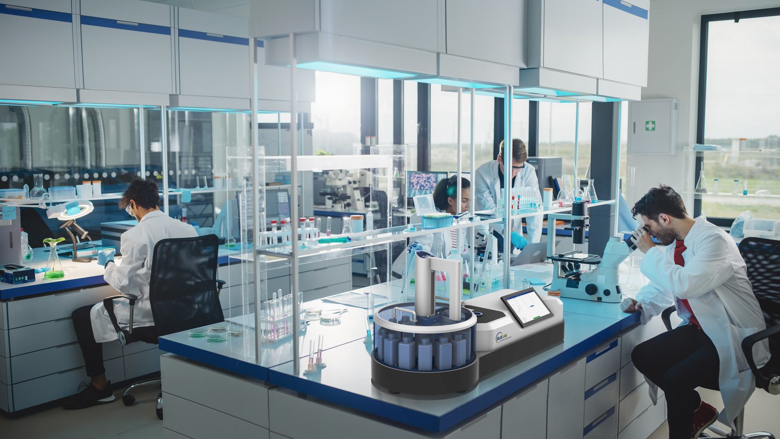 BlueLab-Analysegerät mit bestücktem Probenkarussell auf einem Labortisch, im Hintergrund arbeitendes Laborpersonal